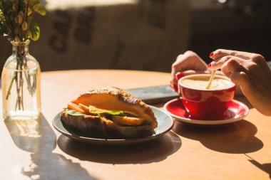 Bir kişi bir fincan kahve ve bir kruvasanla bir masada oturuyor. Kahve fincanının yanındaki masada bir cep telefonu var.