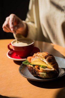 Bir kişi bir fincan kahve ve bir kruvasanla bir masada oturuyor. Kahve fincanının yanındaki masada bir cep telefonu var.