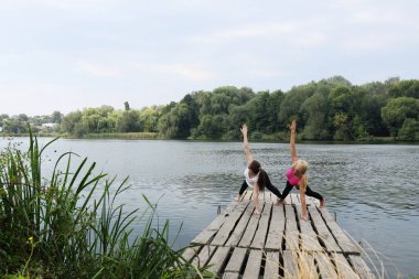 İki genç kız, nehrin kıyısındaki ahşap iskelede yoga dengesi egzersizi yapıyor. Gençlerin fiziksel ve zihinsel sağlığı kavramı