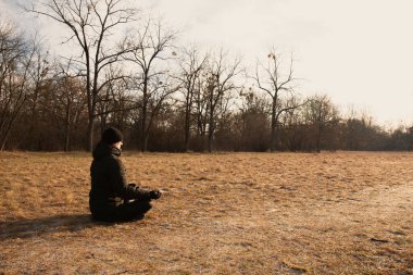 Bir adam soğuk mevsimde bir tarlada kuru çimenlerde oturur ve lotus pozisyonunda meditasyon yapar. Adam siyah bir ceket ve siyah bir şapka giyiyor.