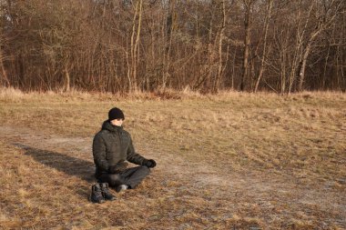 Bir adam soğuk mevsimde bir tarlada kuru çimenlerde oturur ve lotus pozisyonunda meditasyon yapar. Adam siyah bir ceket ve siyah bir şapka giyiyor, ayakkabıları yakınlarda.