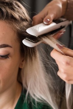 Bir stilist bir kadının saçına hacim katmak için saç maşası kullanır. Yakın plan.