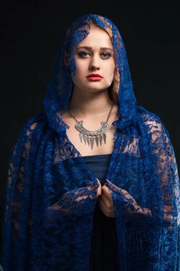 Mavi elbiseli ve kapüşonlu mavi dantel pelerinli bir kadın. Gümüş bir Scythian kolyesi ve kırmızı bir ruj sürüyor.