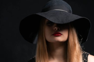 Siyah şapkalı ve kırmızı rujlu bir kadın. Yüzünün gizemli ifadesi şapkanın geniş kenarlarıyla kaplıdır.