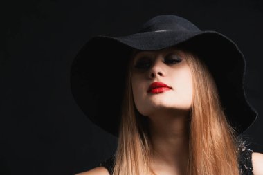 Siyah şapkalı ve kırmızı rujlu bir kadın. Yüzünün gizemli ifadesi şapkanın geniş kenarlarıyla kaplıdır.