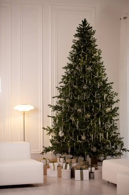 Işıkları ve süsleri olan büyük bir Noel ağacı beyaz duvarları ve beyaz bir sandalyesi olan bir odada oturur.