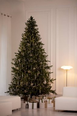 Işıkları ve süsleri olan büyük bir Noel ağacı beyaz duvarları ve beyaz bir sandalyesi olan bir odada oturur.