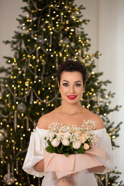 Bir kadın elinde bir buket çiçekle bir Noel ağacının önünde duruyor. Sahne şenlikli ve neşeli, kadının kırmızı ruju resme renk katıyor.