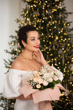 Bir kadın elinde bir buket çiçekle bir Noel ağacının önünde duruyor. Sahne şenlikli ve neşeli, kadının kırmızı ruju resme renk katıyor.