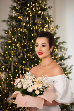 Bir kadın elinde bir buket çiçekle bir Noel ağacının önünde duruyor. Sahne şenlikli ve neşeli, kadının kırmızı ruju resme renk katıyor.
