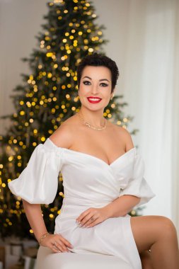 Beyaz elbiseli bir kadın Noel ağacının önündeki beyaz bir kanepede oturuyor. O kırmızı bir kolye ve bir yüzük takıyor. Sahne şenlikli ve ciddidir.