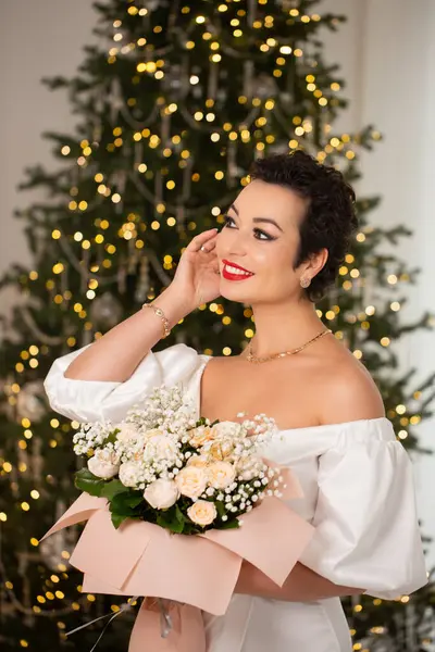 Bir kadın elinde bir buket çiçekle bir Noel ağacının önünde duruyor. Sahne şenlikli ve neşeli, kadının kırmızı ruju resme renk katıyor.