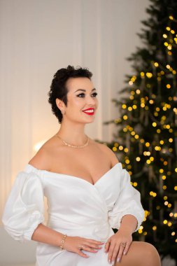 Beyaz elbiseli bir kadın Noel ağacının önünde oturuyor. Kırmızı bir kolye ve kırmızı bir yüzük takıyor. Sahne neşeli ve neşeli, kadın mutlu ve mutlu görünüyor.