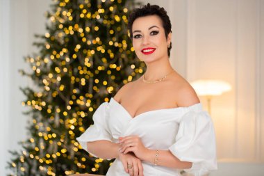 Beyaz elbiseli bir kadın Noel ağacının önündeki beyaz bir kanepede oturuyor. O kırmızı bir kolye ve bir yüzük takıyor. Sahne şenlikli ve ciddidir.