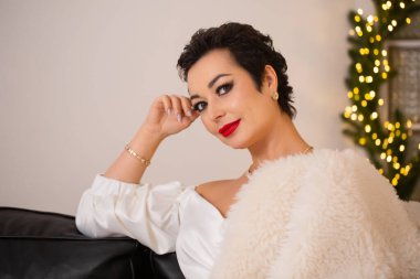 Omzunda beyaz kürklü bir kürkle bir kadın koltukta oturuyor. O bir elbise ve altın bir bilezik takıyor. Sahne, Noel süsleriyle dolu bir odada geçiyor.