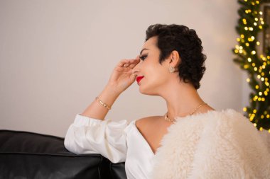 Omzunda beyaz kürklü bir kürkle bir kadın koltukta oturuyor. O bir elbise ve altın bir bilezik takıyor. Sahne, Noel süsleriyle dolu bir odada geçiyor.