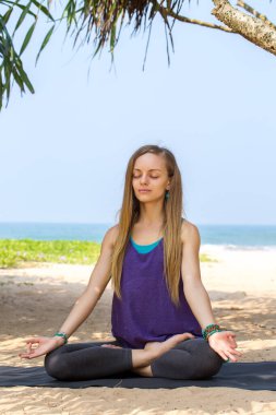 Bir kadın sahilde oturuyor, bir palmiye ağacının altında ve okyanus kenarında meditasyon yapıyor. Sahne huzurlu ve rahatlatıcı.