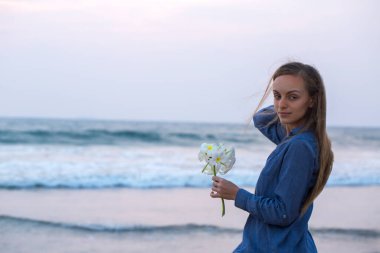 Bir kadın elinde beyaz su tesisatı çiçekleriyle gün batımına karşı okyanusun kenarında duruyor. Sahne huzurlu ve huzurlu.