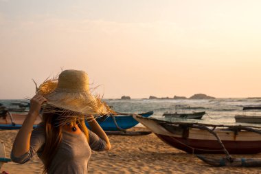 Hasır şapkalı bir kadın sahilde teknelerin yanında duruyor. Gökyüzü mavi ve turuncunun karışımıdır, sıcak ve davetkar bir atmosfer yaratır.