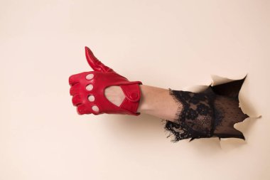 Kırmızı deri eldivenli bir kadının eli iyi bir jest gösteriyor. El, bej kağıttaki bir delikten itilir..