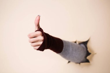 Örülmüş bordo eldivenli bir kadının eli bir jest gösterir. El, bej kağıttaki bir delikten itilir..