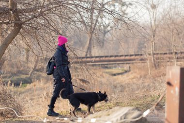 Şapkalı ve ceketli bir kadın parkta tasmalı bir köpekle yürüyor.