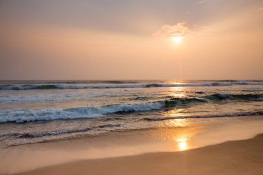 Güneş okyanusun üzerinde batıyor, kumun üzerine sıcak bir parıltı saçıyor. Su sakindir ve gökyüzü turuncu ve pembe renklerin bir karışımıdır. Sahne huzurlu ve huzurlu.