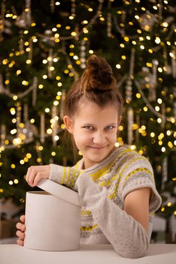 Teenage girl in knitted sweater smiling slyly looking at camera and opening gift near decorated Christmas tree clipart