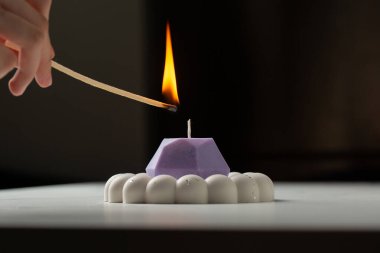 A person lights a purple candle with a match. The candle is on a white plaster stand. clipart