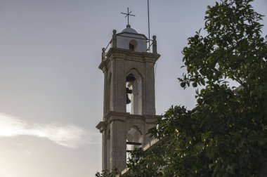 Huzurlu çan kulesi ve bir kilisenin haçı, yemyeşil yeşillikler çerçeveli yumuşak bir günbatımı gökyüzüne karşı. Lania köyü. Limasol İlçesi, Kıbrıs