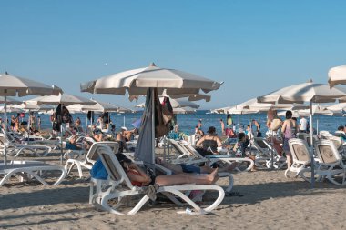 Larnaca, Kıbrıs - 17 Temmuz 2022: Güneşli bir yaz günü Mackenzie Plajı 'nda plaj sandalyelerinde şemsiye altında dinlenen turistler