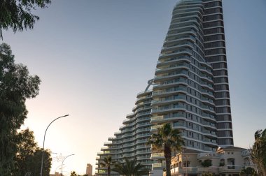 Benzersiz bir tasarıma sahip modern gökdelen binası gün batımında bir şehir caddesinin üzerinde yükseliyor. Limasol, Kıbrıs