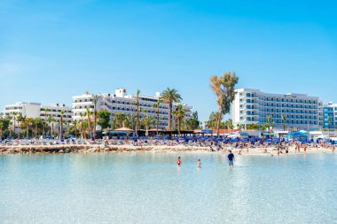 Ayia Napa, Cyprus - July 23, 2018: Tourists are enjoying a sunny day at the beach, swimming in the clear blue water, sunbathing, and relaxing on a beautiful Nissi beach in front of hotels clipart