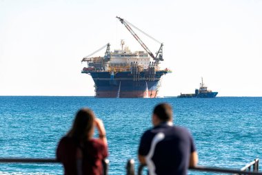 Rıhtımdan büyük bir açık deniz konaklama platformunu gözlemleyen turistler, yakınlarda küçük bir gemi seyrediyor.