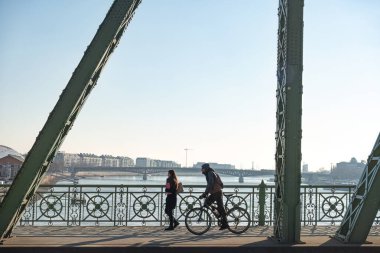 Budapeşte, Macaristan - 07 Ocak 2019: Bisiklet süren genç bir adam ve Budapeşte, Macaristan 'daki ünlü Özgürlük Köprüsü üzerinde yürüyen genç bir kadın