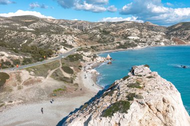 Tourists walking on aphrodite's rock beach with turquoise water and a highway in the background on a sunny day. Paphos District clipart