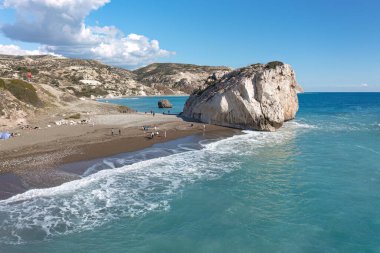 Tourists enjoying the beach and the view of the sea stack called Aphrodite's rock (Petra tou Romiou) on a sunny day. Paphos District, Cyprus clipart