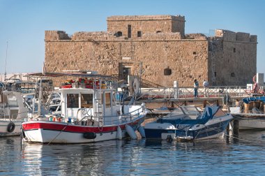 Açık mavi gökyüzünün altında tarihi Paphos şatosuyla dolu hareketli bir limana demirlemiş tekneler, tarih ve deniz yaşamının bir karışımını yakalıyor. Kıbrıs