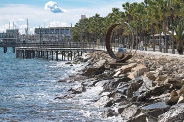 Limasol, Kıbrıs - 02 Ocak 2024: Metalik, modern bir sanat heykeli tarafından kayalara çarpan dalgalar, limasol kıyılarında, cythe people walking by