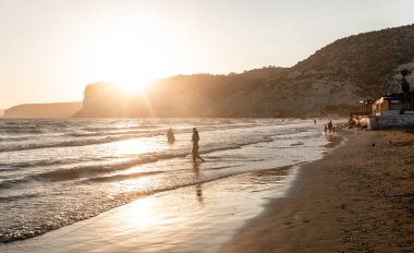 Günbatımında Kourion sahilinde yürüyen turistler deniz suyuna yansıyan güneşle