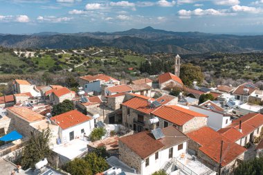 Kıbrıs Rum Kesimi 'nin Larnaca ilçesindeki Pano Lefkara köyünün hava manzarasında geleneksel mimari, kırmızı kiremitli çatılar ve arkasında Troodos Dağları olan bir kilise sergileniyor.
