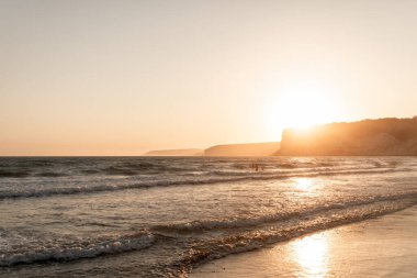Kourion Sahili 'nde altın gün batımı ışığı ve dalgalar kıyıya vuruyor, insanlar uzaklarda yüzüyor. Episkopi, Limasol İlçesi, Kıbrıs
