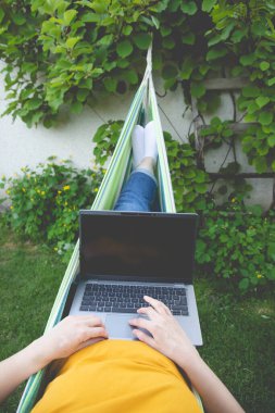 Genç ve güzel bir kadın bir hamakta dizüstü bilgisayarla bahçede yatıyor.. 