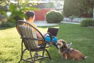 Güneşli bir günde avluda tüylü kahverengi köpeğinin yanında dizüstü bilgisayarda çalışan bir kadın. Evden çalışan serbest çalışan bir iş kadını..