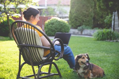 Güneşli bir günde avluda tüylü kahverengi köpeğinin yanında dizüstü bilgisayarda çalışan bir kadın. Evden çalışan serbest çalışan bir iş kadını..
