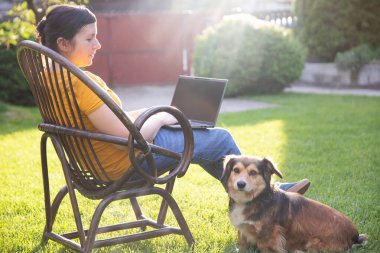 Güneşli bir günde avluda tüylü kahverengi köpeğinin yanında dizüstü bilgisayarda çalışan bir kadın. Evden çalışan serbest çalışan bir iş kadını..