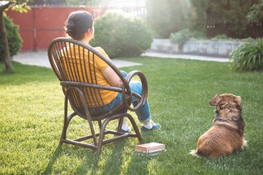 Kadın kitap okuyor, güneşli bir günde tüylü kahverengi köpeğiyle sandalyede dinleniyor. Hayvanlarla bağ kurma ve birlikte olmaktan keyif alma kavramı.