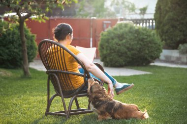 Kadın kitap okuyor, güneşli bir günde tüylü kahverengi köpeğiyle sandalyede dinleniyor. Hayvanlarla bağ kurma ve birlikte olmaktan keyif alma kavramı.