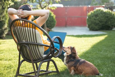 Güneşli bir günde avluda tüylü kahverengi köpeğinin yanında dizüstü bilgisayarda çalışan bir kadın. Evden çalışan serbest çalışan bir iş kadını..