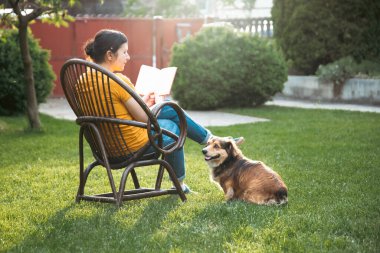 Kadın kitap okuyor, güneşli bir günde tüylü kahverengi köpeğiyle sandalyede dinleniyor. Hayvanlarla bağ kurma ve birlikte olmaktan keyif alma kavramı.